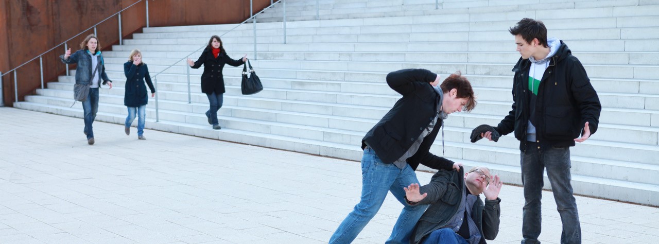 Einem am Boden liegender Mann werden Faustschläge von zwei Jugendlichen Männern angedroht. Drei Frauen eilen zur Hilfe.