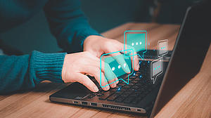 Eine Person sitzt am Tisch, man sieht ihre Hände, sie hat vor sich einen Laptop und tippt darauf. Über der Tastatur erscheinen futuristisch animierte Graphen und Charts.