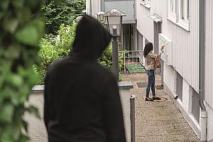 Person in schwarzer Kapuzenjacke beobachtet eine Frau, die den Briefkasten öffnet.