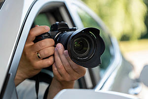A paparazzi sits in his car and takes pictures of a famous person. Spy with camera in the car. A private detective, sitting inside the car, taking pictures with the camera. Eine Person sitzt im Auto und macht Fotos mit der Kamera.