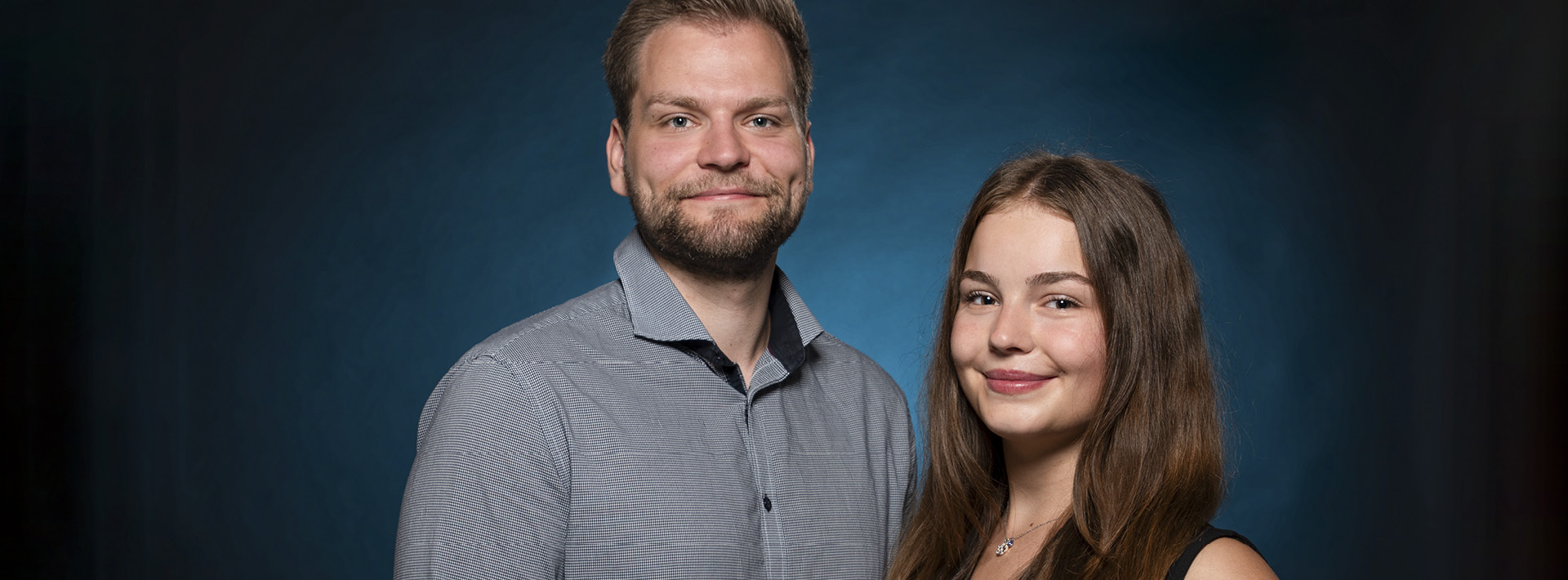 ein junger Mann und eine junge Frau blicken Frontal in die Kamera. Sie stehen vor einem dunkelblauem Hintergrund